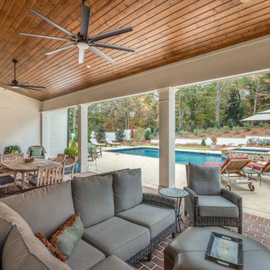 Outdoor patio with grey cushioned furniture under a wooden ceiling featuring fans. A pool is visible outside, surrounded by lounge chairs and a table with umbrella. The area is enclosed by a white brick wall and trees in the background.