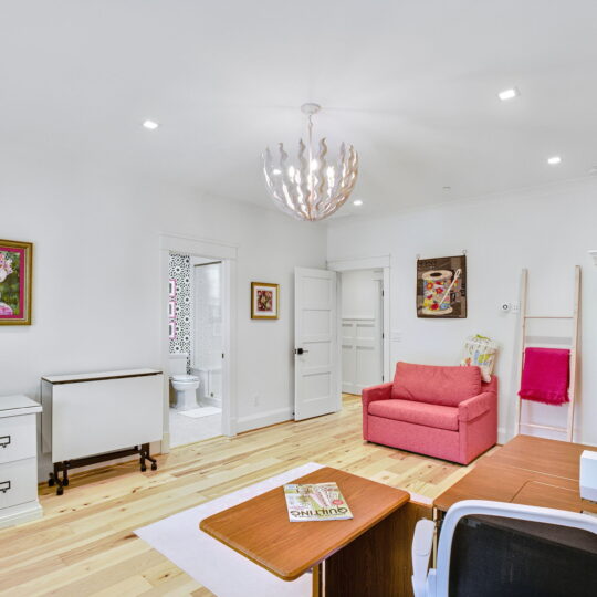 A bright sewing room with wooden floors features a sewing machine on a desk, a pink cushioned chair, a cupboard, and a unique ceiling light. Artwork adorns the walls, and a doorway leads to a bathroom. Shelves hold various supplies.