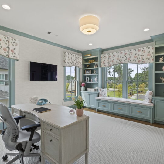 Home office with a large wooden desk and ergonomic chair. Built-in teal shelves and window seat with floral curtains. Windows offer views of greenery and houses. Neutral walls and carpet, modern ceiling light, and a wall-mounted TV add to the decor.