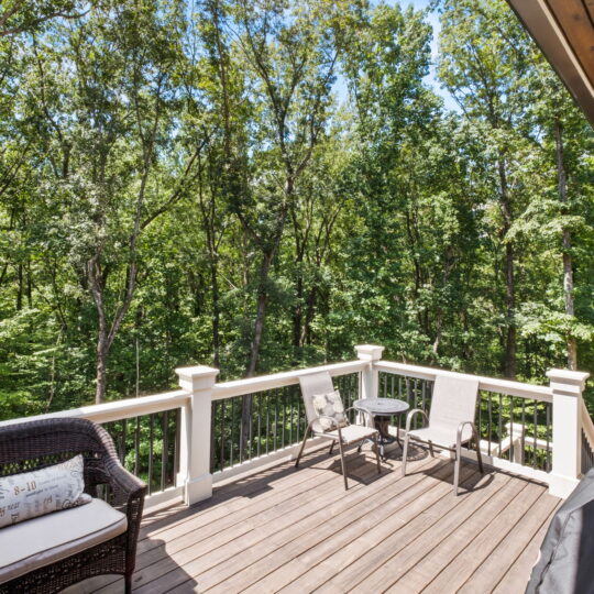 A wooden deck with a black wicker sofa and two beige lounge chairs around a small circular table, overlooking a lush, green forest. The sky is clear and sunny, casting bright light onto the scene.