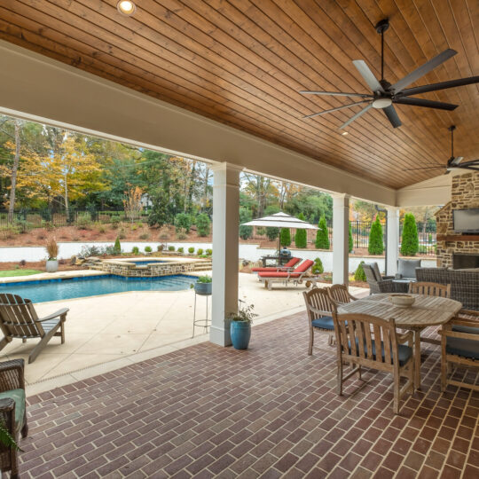 Covered patio with brick flooring featuring wooden dining furniture, outdoor seating, and a ceiling fan. Overlooking a pool area with lounge chairs and an umbrella. Surrounded by trees and greenery.