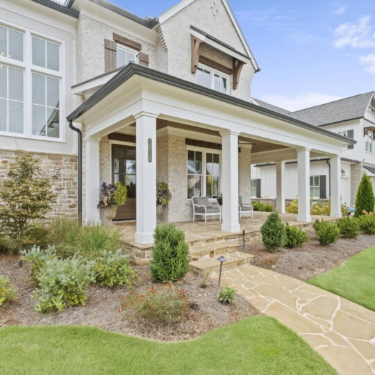 A spacious two-story house with a stone facade and white pillars, featuring a covered porch with seating. The front yard is landscaped with shrubs and a stone pathway. Another similar house is visible in the background under a partly cloudy sky.