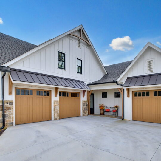 A modern house with white siding and stone accents features three brown wooden garage doors. The garage roof has dark metal accents. An American flag is displayed to the left, and a clear blue sky is in the background.