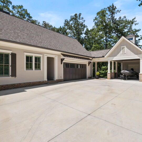 A spacious driveway leads to a two-car garage attached to a modern house with light-colored siding and brick accents. The garage door is closed, and theres an open carport to the right housing a covered vehicle. Trees and a blue sky are in the background.