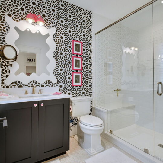 Modern bathroom with black and white patterned wallpaper, black vanity with double sink, and large white-framed mirror. Glass shower enclosure on the right. Three small red-framed mirrors hang vertically above the toilet. White rugs on tiled floor.