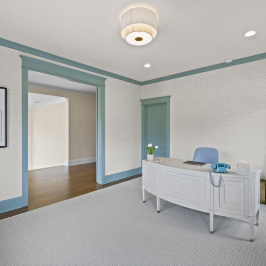 A home office with a white desk and a blue chair on a gray carpet. A vintage blue telephone sits on the desk. Light blue trim accents the walls and door. A shelf holds various decor items. A framed artwork hangs on the wall. Ceiling light fixture above.