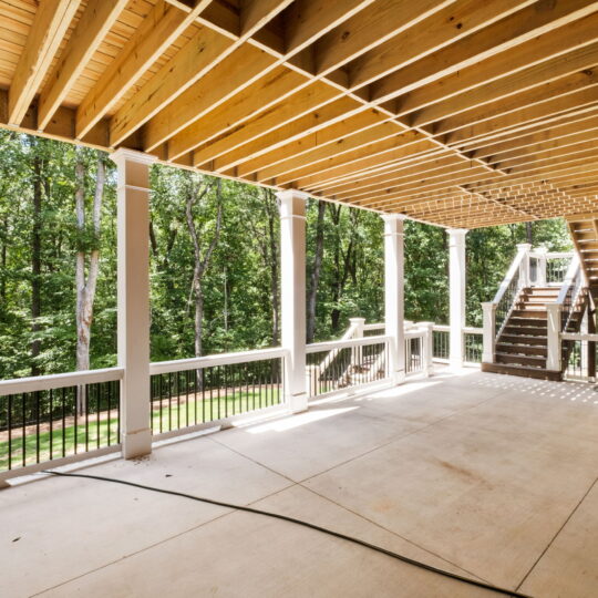 Covered patio with wooden ceiling beams and brick wall, overlooking a lush, green forest. A set of wooden stairs leads down from the deck. A garden hose is coiled on the smooth concrete floor. White railings line the space.