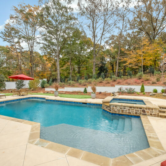 A backyard pool surrounded by a tiled patio with lounge chairs and a red umbrella. The pool has a small stone waterfall feature. Trees with autumn foliage are in the background under a partly cloudy sky.