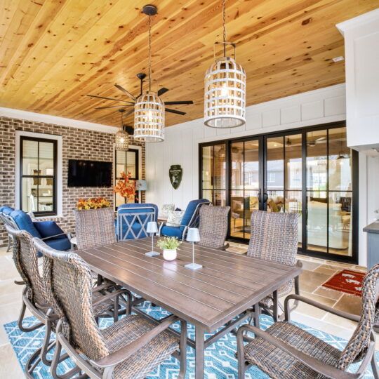 A covered patio features a wooden ceiling, wicker chairs around a rectangular table, and two hanging lamps. A TV is mounted on a brick wall. Large glass doors lead to another room, and a rug adds a pop of color under the table.