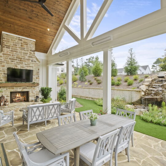 Elegant patio with stone fireplace, wooden ceiling, and TV. Outdoor dining set and chairs arranged on a stone floor. Greenery and a waterfall feature in the background, under a bright, open sky.