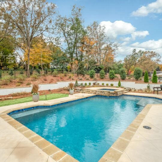 A backyard features a rectangular swimming pool surrounded by a stone patio. There are lounge chairs, an outdoor table with an umbrella, and a grill. Trees with autumn foliage and a clear blue sky are visible in the background.