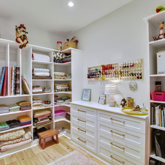 A neatly organized craft room with shelves holding fabric, thread spools, and books. A white dresser with gold handles is in the center, topped with a lamp and decorative items. A small wooden stool is on the floor, and colorful rugs add warmth.