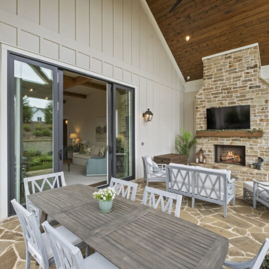 Outdoor patio with a wooden dining table and chairs, facing a stone fireplace with a mounted TV. Cozy seating area includes cushioned chairs and a sofa. Sliding glass doors open to a living room inside. Lush greenery decorates the space.