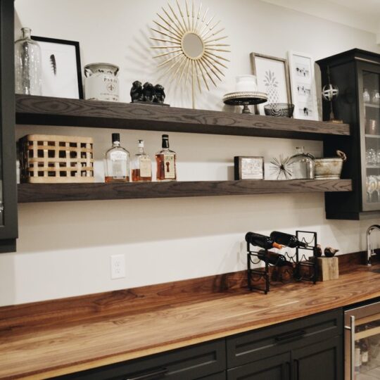 A cozy home bar with dark cabinets and wooden countertop. Shelves display glassware, bottles, and decorative items. A wine rack and small fridge are beneath. A sunburst mirror and framed art hang on the wall.