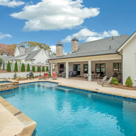 A backyard with a rectangular pool and hot tub surrounded by a stone deck. There is a covered patio area with outdoor furniture and a grill. The house has white brick and multiple chimneys. The sky is blue with scattered clouds.