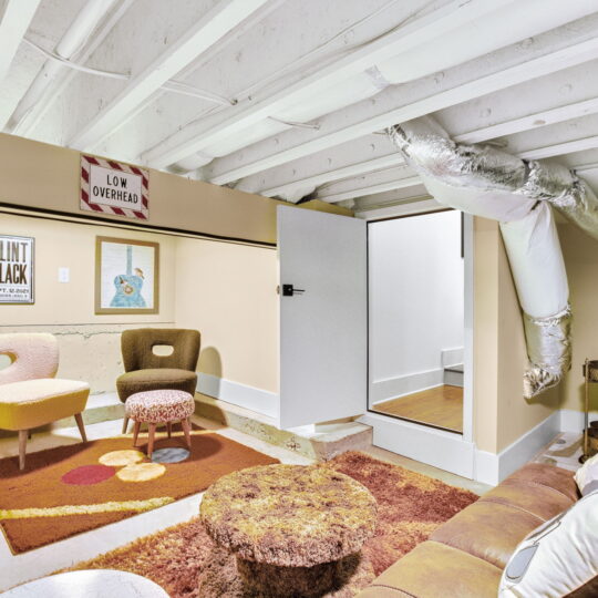 A cozy basement room with a brown couch, colorful chairs, and a shaggy rug. Wall decor includes framed pictures and a Low Overhead sign. Exposed ductwork and beams enhance the rustic atmosphere, and a small step leads to a higher level.