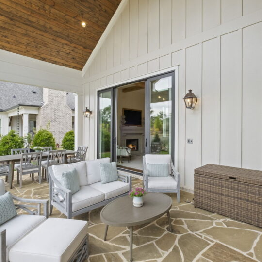 Modern outdoor patio with white cushioned seating, light gray chairs, and a round coffee table on a stone floor. A wicker storage box is near a sliding door, beyond which is an interior with a fireplace. Dining area and greenery in the background.