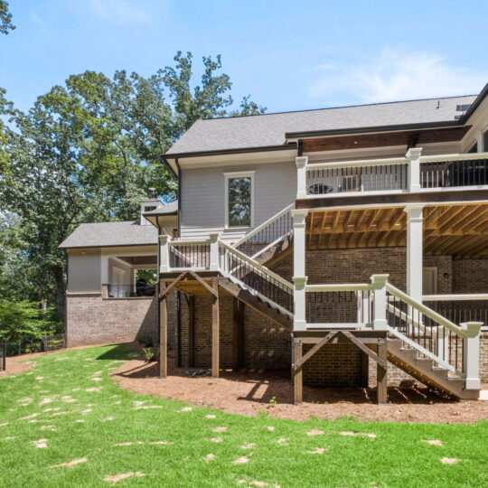 Backyard view of a large brick house with a wooden staircase leading to a second-floor deck. The area features a well-maintained lawn, trees, and a fenced section. The house has multiple levels and a covered patio underneath the deck.