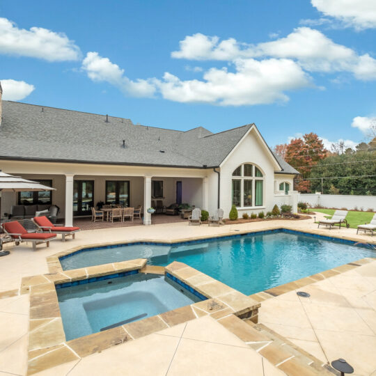 Luxurious backyard with a stone patio featuring a large swimming pool, hot tub, and sun loungers under umbrellas. The house has a modern design with arched windows. Lush greenery and a clear blue sky with a few clouds are in the background.