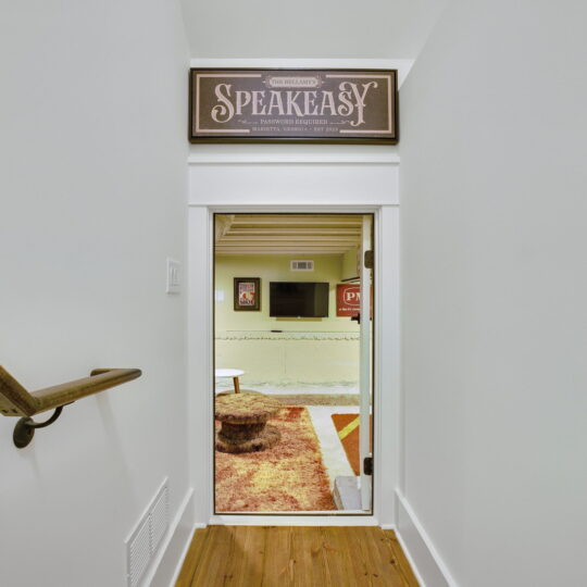 A narrow hallway with a wooden floor and a handrail leads to a small door with a Speakeasy sign above it. Beyond the door, a room with a vintage-themed interior and a round table is visible.