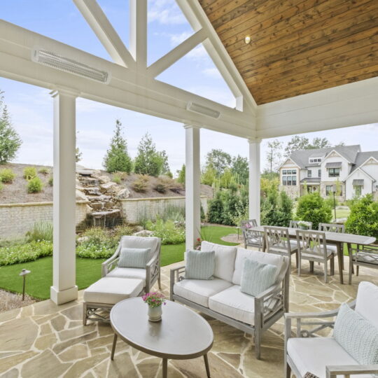 Patio with white cushioned furniture under a wooden ceiling. Overlooks a stone patio with a table, chairs, and a waterfall feature in a landscaped backyard. Houses and greenery are visible in the background under a blue sky.