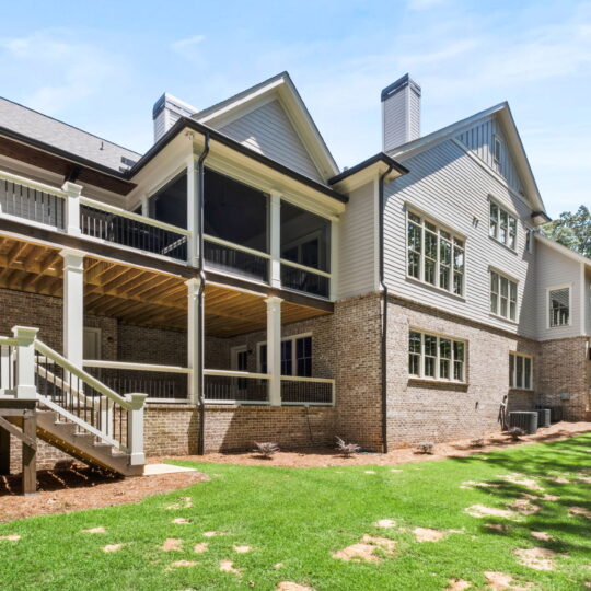 Spacious two-story house with a brick and white siding exterior, featuring multiple covered balconies and large windows. The house overlooks a grassy yard with surrounding trees under a clear blue sky.