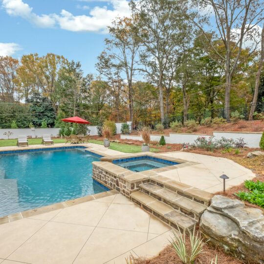 A backyard features a large swimming pool surrounded by stone and tile decking. Lounge chairs and an umbrella are on a patio, with trees and a white fence in the background. The scene is serene, with clear skies above.