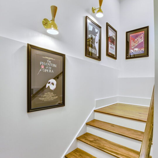 Staircase with wooden steps and white walls, decorated with framed posters of musicals including The Phantom of the Opera and others. Wall-mounted lights illuminate the space.