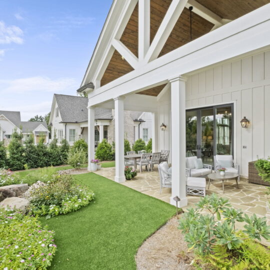 A spacious patio with white siding and stone accents, featuring outdoor furniture, potted plants, and a small garden. The patio overlooks a manicured lawn and other houses in the background are visible under a partly cloudy sky.