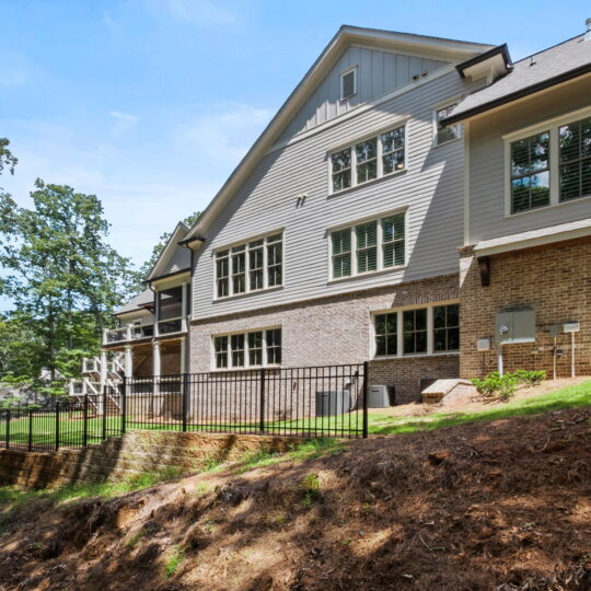 The image shows the back of a modern, multi-story house with brick and siding exterior. It features large windows, multiple levels, and a fenced patio. The surrounding area is landscaped with trees and a grassy slope. The sky is clear and sunny.