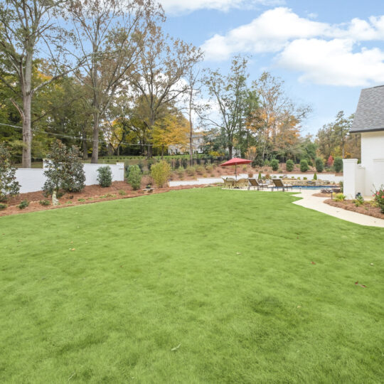 A scenic backyard featuring a large green lawn bordered by flowerbeds and a white fence. A curved pathway leads to a patio with a seating area beneath a red umbrella. Trees and bushes line the fence, with a house partially visible on the right.