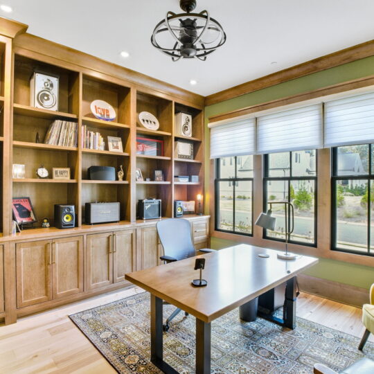 Home office with wooden shelves filled with books, records, and audio equipment. A desk with a black chair faces a large window with white blinds. Two yellow armchairs and a patterned rug add color, and a modern light fixture hangs above.