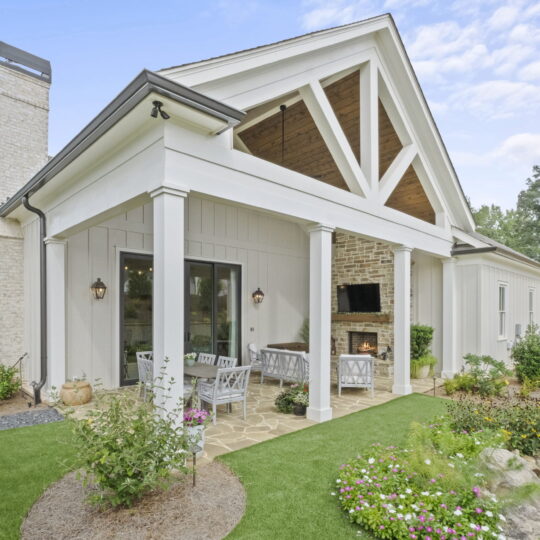 A modern white house with a peaked roof and large covered patio. The patio has outdoor seating and a fireplace. The lawn is landscaped with flowers and rocks. The sky is clear and trees are visible in the background.