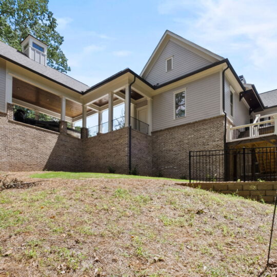 A spacious, two-story home with gray siding and brick foundation. It features multiple gabled roofs and a covered porch. The backyard has a sloped lawn, trees in the background, and a black metal fence on a stone retaining wall.