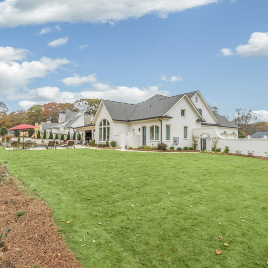 A large, elegant white house with steep gabled roofs and arched windows, set amidst a vast, neatly manicured green lawn. The backyard features a patio with outdoor furniture and a red umbrella. The sky is partly cloudy, and trees are visible in the background.