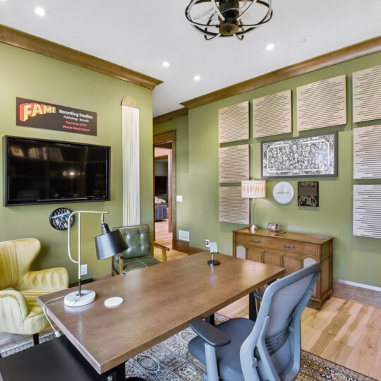 A modern office with a large wooden desk, green and yellow chairs, and a wall-mounted TV. The walls are olive green, adorned with sound panels and a framed print. Ceiling lights and a patterned rug complete the space.