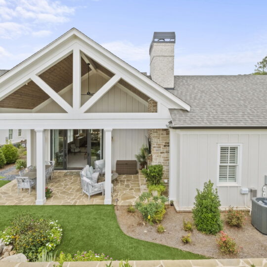 A modern house with white siding and large gable roof, featuring a covered patio with outdoor seating and a stone floor. The backyard is landscaped with small bushes, flowering plants, and a patch of artificial grass.