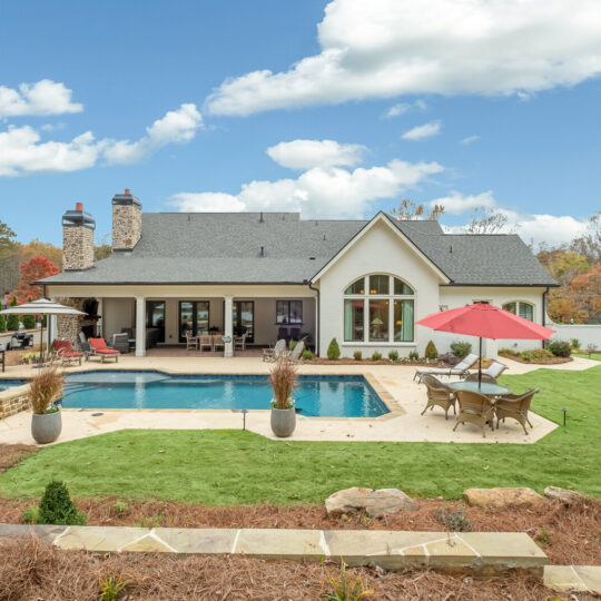 A spacious backyard with a rectangular pool surrounded by a stone patio. Theres a large house with multiple windows in the background. Outdoor furniture, including a table with a red umbrella, is arranged on the patio. The sky is partly cloudy.