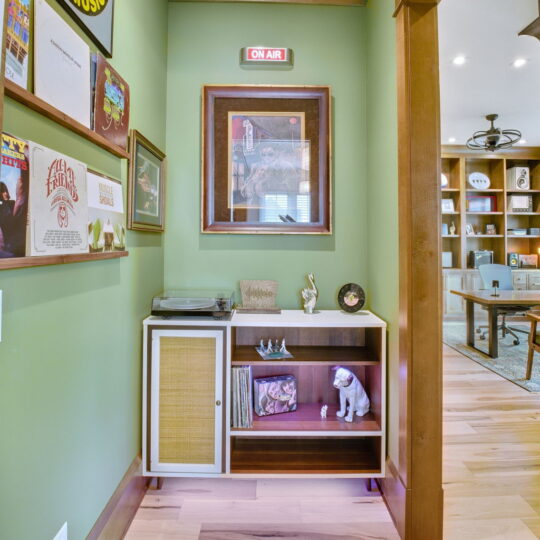 A cozy corner with a green accent wall features vinyl records and framed art. A shelf holds a vintage turntable, decorative items, and a small dog sculpture. A hallway leads to a room with bookshelves and a ceiling fan visible in the background.