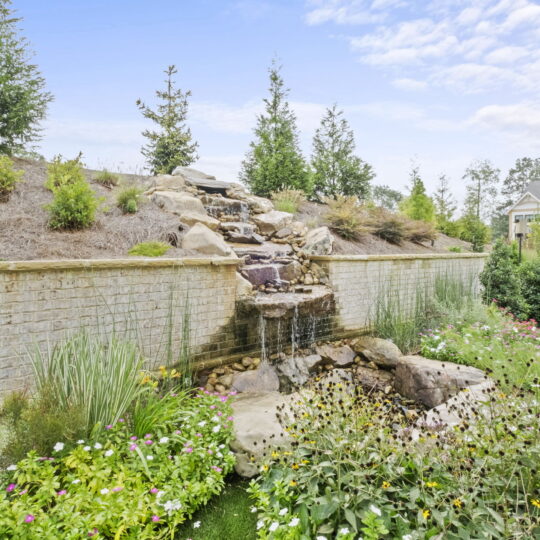 A garden with a small waterfall cascading over rocks, surrounded by green plants and flowers. A brick wall supports the waterfall, and trees and shrubs are visible in the background. A house is partially seen to the right.