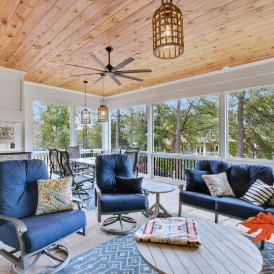 Spacious sunroom with a wooden ceiling, large windows, and blue cushioned chairs around round tables. A grill is in the corner. Ceiling fans and hanging lights add ambiance. Autumn leaves decorate one table.