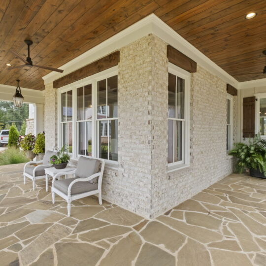 Covered porch with light stone flooring and a wooden ceiling. Two gray cushioned chairs with a small table are set near large windows. Ceiling fans are mounted above, and potted ferns sit by the houses entrance. A driveway and garden are visible.