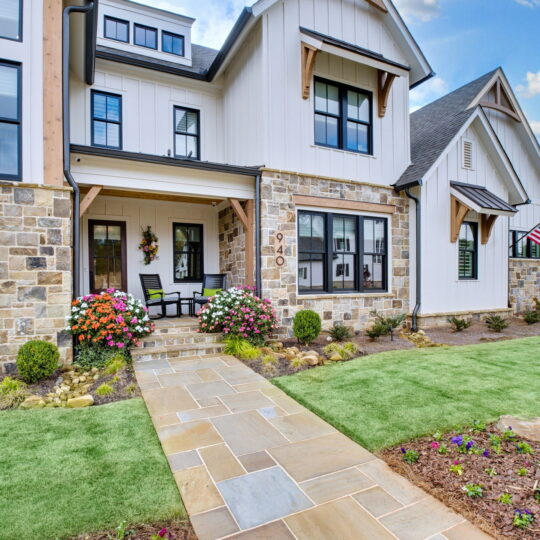 A modern two-story house with a stone and white siding exterior. The front porch has rocking chairs and colorful flowers. A paved walkway leads through green grass, and an American flag is displayed. The sky is clear with a few clouds.