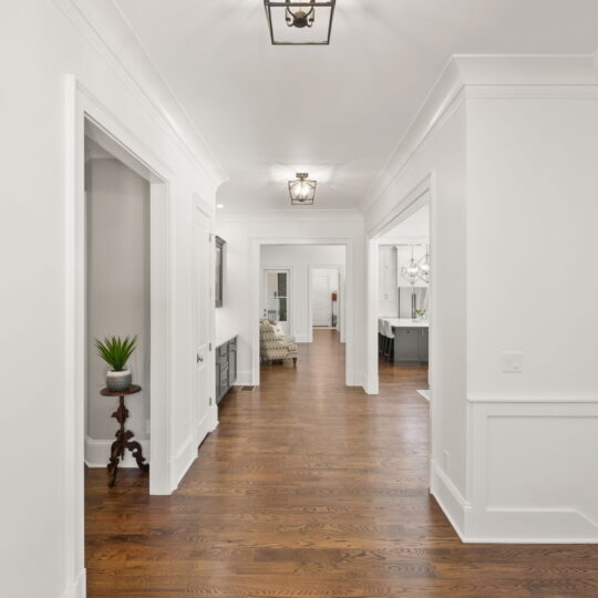 Bright, elegant hallway with hardwood floors and white walls. Room with a plant on a side table to the left, framed pictures along the corridor, and a glimpse into a well-lit living area at the end. Ceiling lights enhance the classic decor.