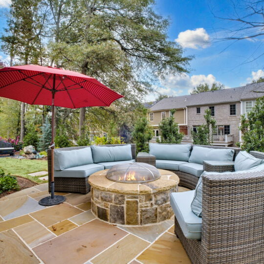 Outdoor patio scene with a circular wicker sofa and light blue cushions around a stone fire pit. A red umbrella provides shade. The background features green trees, shrubs, and brick houses under a blue sky with some clouds.