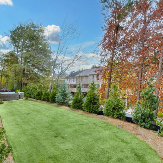 A backyard with a well-maintained grassy area bordered by trees. On the left, a white house is visible, while the background shows neighboring homes and a clear blue sky with scattered clouds.