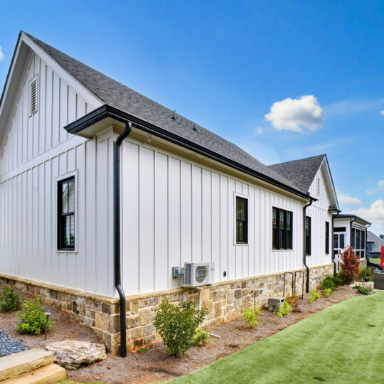 Side view of a modern white house with a peaked roof and black trim. The lower section features stone siding. Theres a gravel path on the left and a grassy area with shrubs on the right, under a clear blue sky with a few clouds.