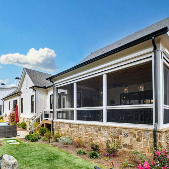 A modern house with white siding and a stone foundation. It features a screened porch, a hot tub, and a landscaped yard with shrubs and flowering plants. The sky is blue with scattered clouds.