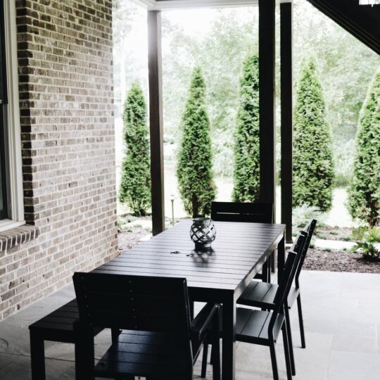 A covered patio with a black table and four chairs on a stone floor. A small decorative object is on the table. Brick wall and neatly trimmed trees in the background. Natural light filters through, creating a calm, inviting atmosphere.