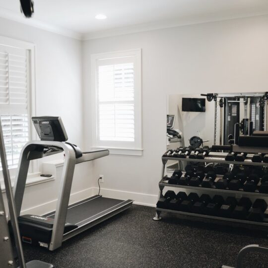 Home gym with a treadmill, a weightlifting station with various dumbbells on a rack, and a wall mirror. The room has white walls, windows with blinds, and a black rubber floor.